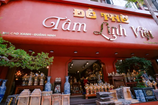 Mặt tiền đỏ Đồ Thờ Tâm Linh Việt với biển chữ lớn, cây xanh, đôi bình xanh trắng và nhiều đồ trưng bày trước cửa.