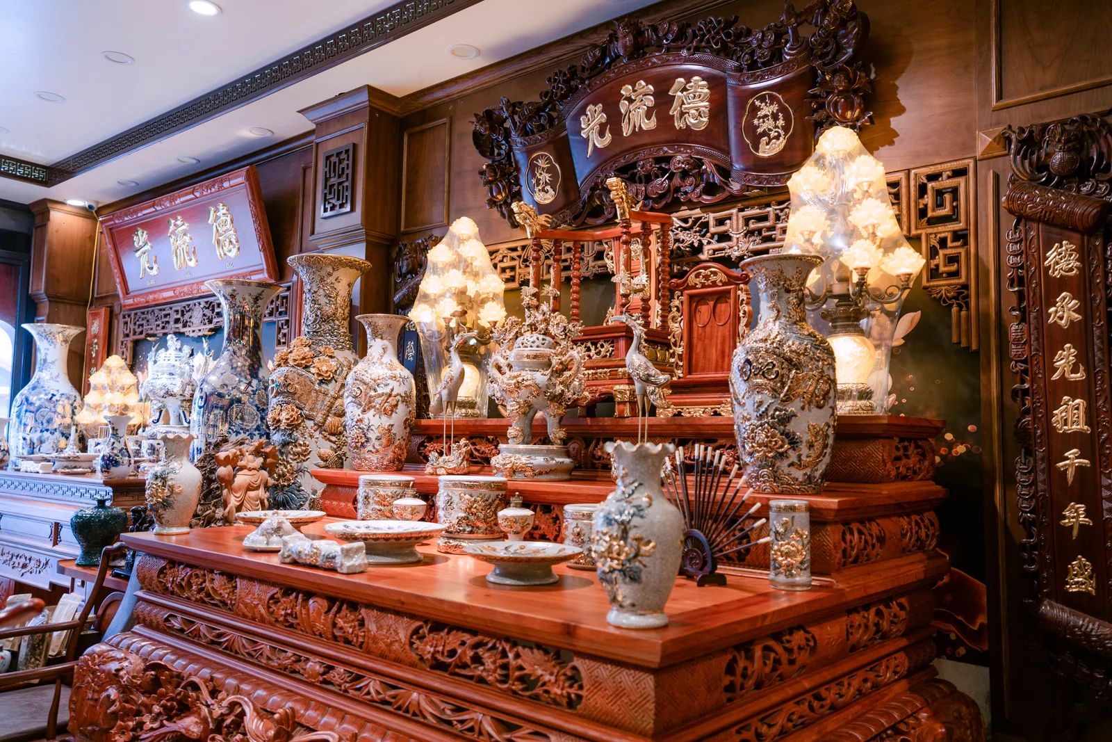 Góc chéo bàn thờ nhiều tầng với mặt bàn chạm dày, bình họa tiết nổi, đỉnh, hạc, đèn và cuốn thư phía trên.