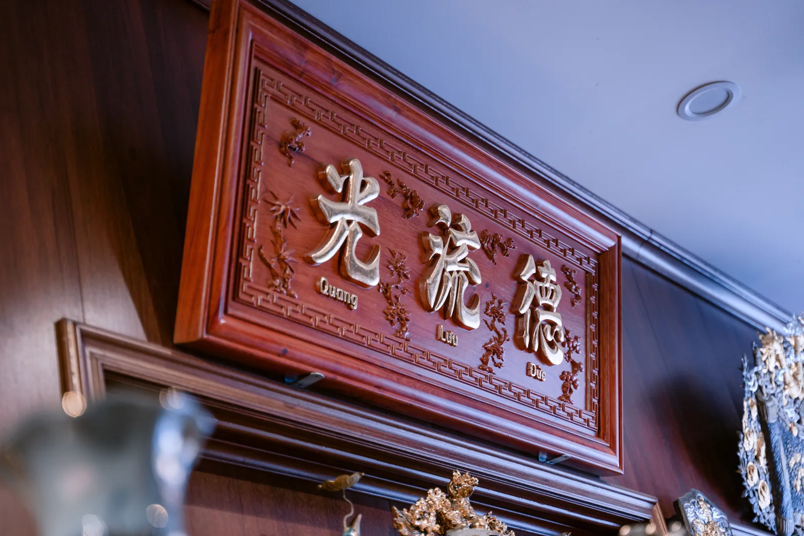 Hoành phi chữ nhật màu nâu đỏ treo cao, ba chữ lớn màu vàng với chữ Quốc ngữ nhỏ bên dưới, viền hình học và hoa văn nhỏ quanh chữ.