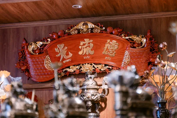 Cuốn thư đỏ vàng lớn với ba chữ nổi và hai đầu cuộn, bao quanh bằng hoa văn; bên dưới có đỉnh, đèn sen và bình cành hoa.