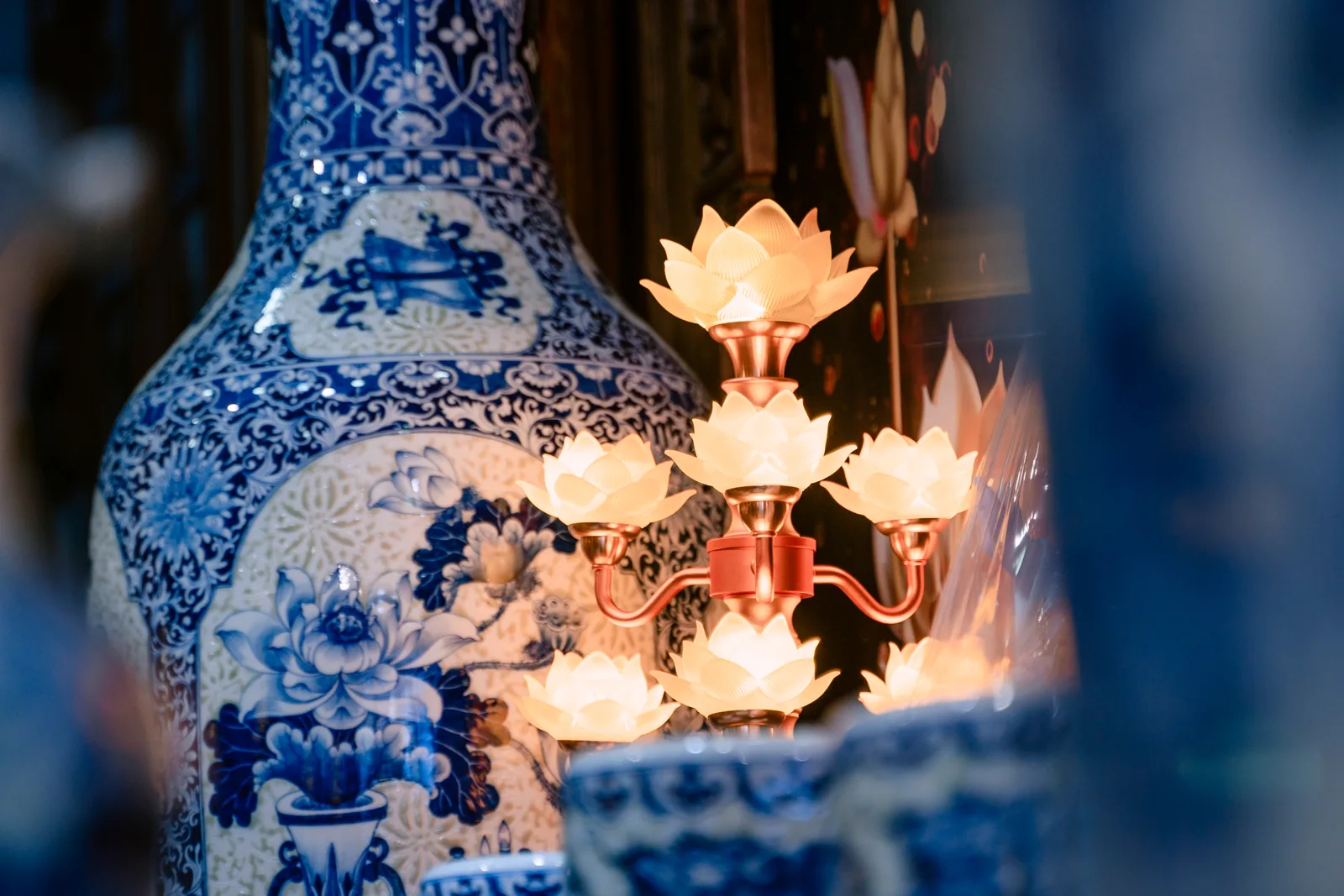 Cụm đèn sen sáng màu vàng ấm với thân và nhánh màu đỏ đồng, đặt cạnh bình lớn xanh trắng có hoa trong một mảng tròn.