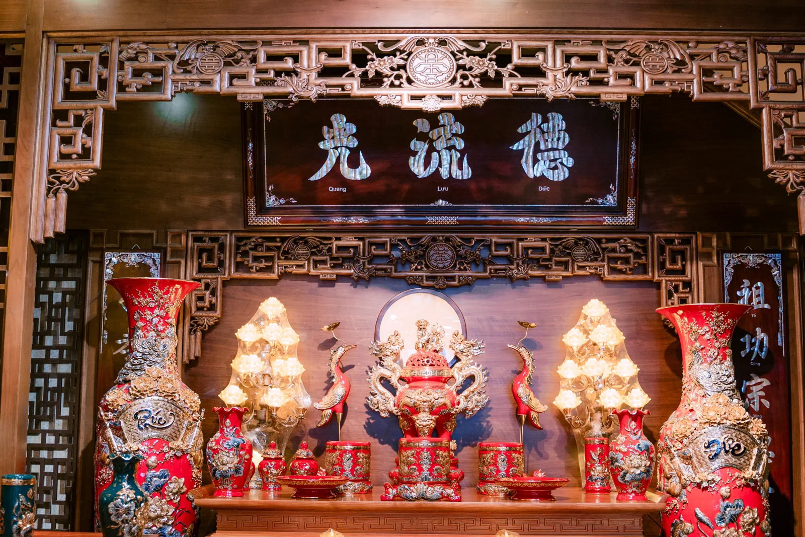 Bàn thờ tông đỏ với đôi bình cao hai bên, đỉnh rồng cùng đôi hạc ở giữa, bát và đồ thấp phía trước, hai đèn sen sáng dưới hoành phi.