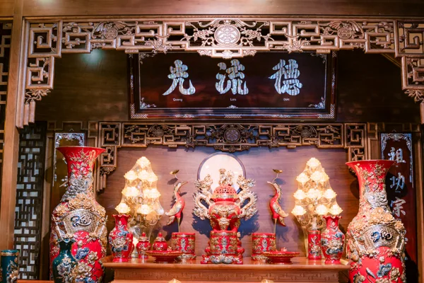 Bàn thờ tông đỏ với đôi bình cao hai bên, đỉnh rồng cùng đôi hạc ở giữa, bát và đồ thấp phía trước, hai đèn sen sáng dưới hoành phi.