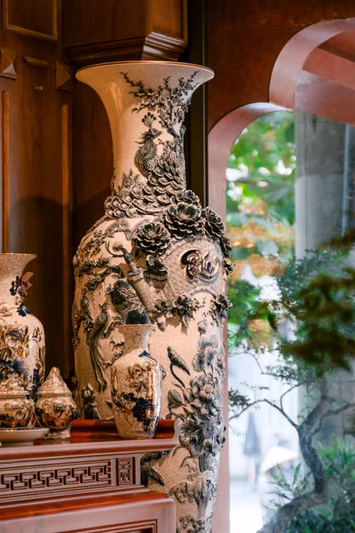 Bình cao nền sáng họa tiết nổi màu sẫm đứng cạnh cửa vòm có cây xanh; bàn phía trước mang các bình nhỏ và một món có nắp.