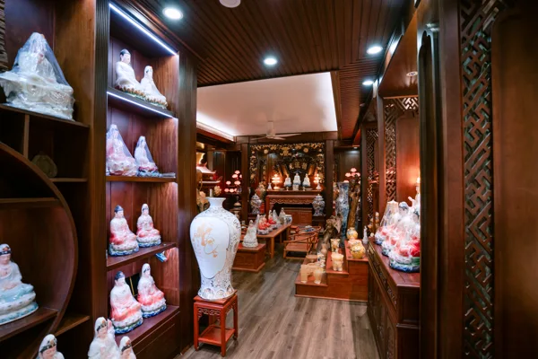 Lối đi trong cửa hàng, kệ tượng nhiều tầng bên trái, bình trắng đặt trên đôn phía trước và vách hoa văn màu nâu bên phải.
