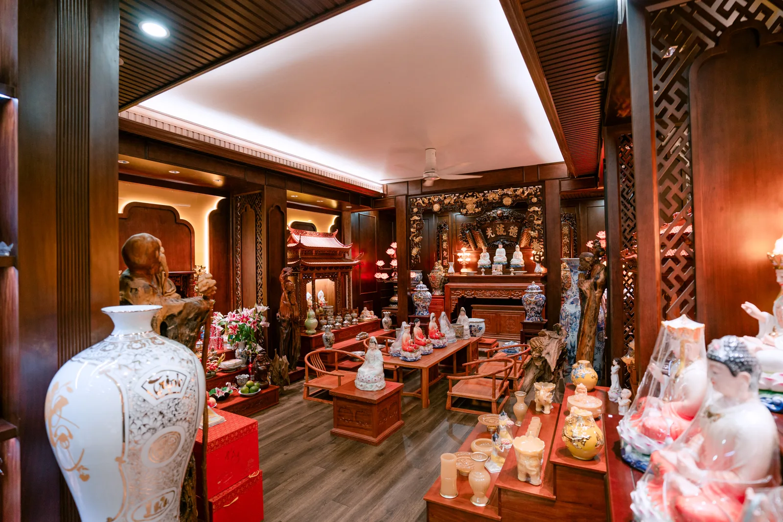 Góc rộng cửa hàng với bình trắng lớn bên trái, bàn ghế ở giữa, nhiều tượng trên bàn và dãy kệ bên phải, bàn thờ ở cuối phòng.
