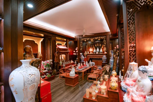 Góc rộng cửa hàng với bình trắng lớn bên trái, bàn ghế ở giữa, nhiều tượng trên bàn và dãy kệ bên phải, bàn thờ ở cuối phòng.