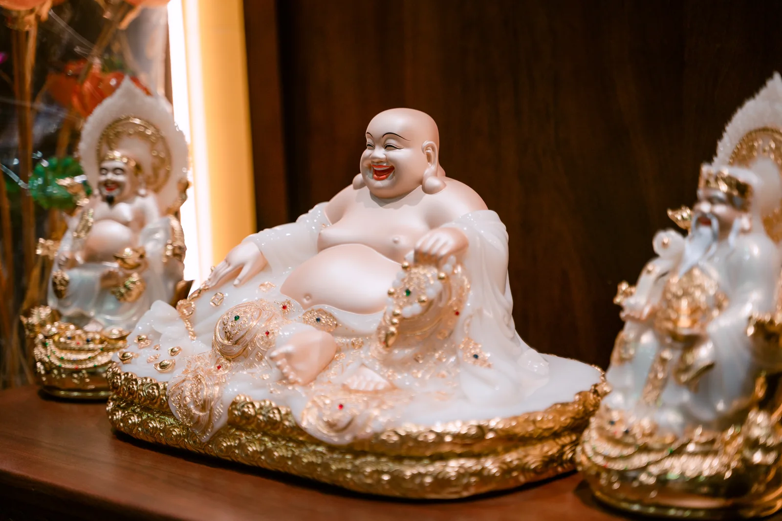 Tượng bụng tròn ngồi cười màu trắng hồng trên bệ rộng nhấn vàng, tay đặt hai bên, hai tượng nhỏ hơn mờ ở mép trái và phải.