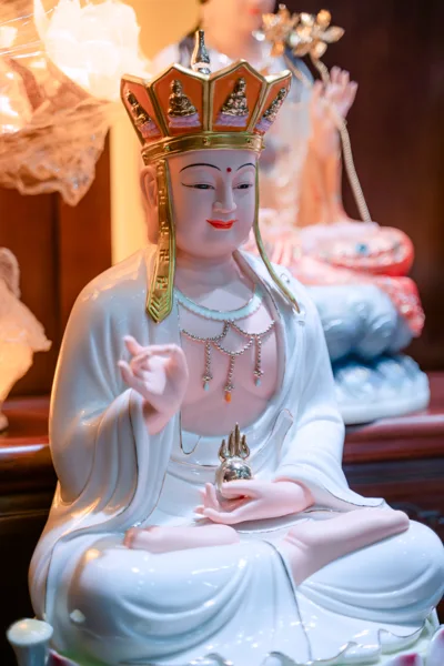 Tượng ngồi áo trắng đội mũ nhiều cánh màu cam viền vàng, một tay giơ lên, tay còn lại đỡ vật nhỏ màu sáng trước bụng.