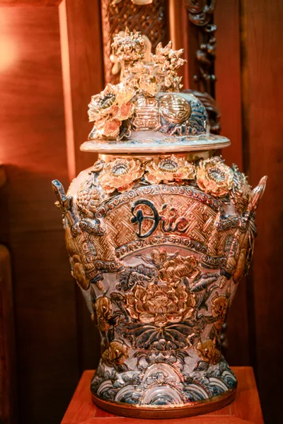 Góc gần chóe chữ Đức có cụm trang trí trên nắp, hàng hoa nổi quanh vai và sen, cá, sóng trên thân; nền gỗ màu nâu đỏ.