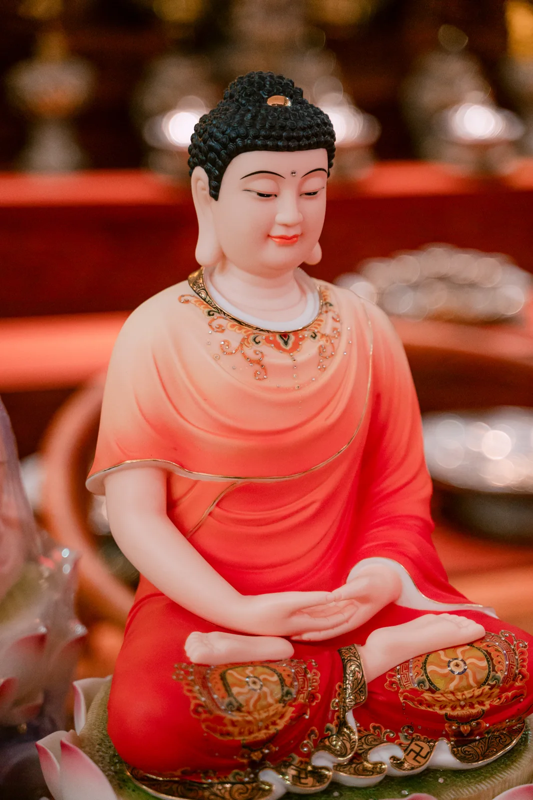 Tượng ngồi tóc xoắn đen, áo chuyển trắng sang đỏ, hai tay xếp giữa lòng, đầu gối trang trí họa tiết tròn màu vàng.