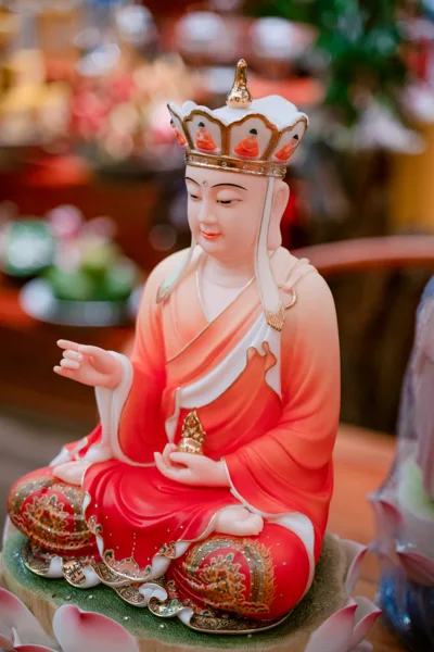 Tượng ngồi áo đỏ trắng đội mũ nhiều cánh viền vàng, một tay đưa ra và một tay giữ vật nhỏ màu vàng trên lòng bàn tay.