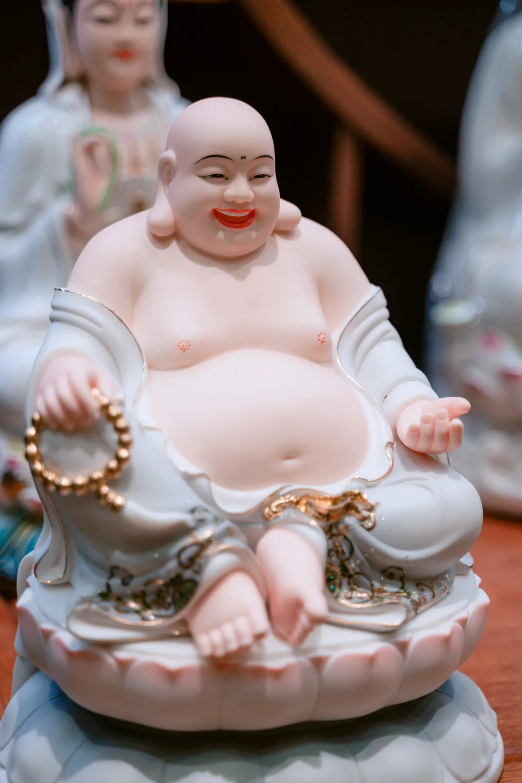 Tượng nhân vật đầu tròn cười rộng, bụng lộ, áo trắng viền vàng, một tay cầm chuỗi hạt và tay kia mở trên đài sen nhạt.