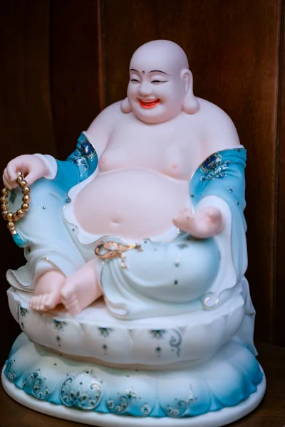 Tượng đầu tròn cười môi đỏ, bụng lộ, áo chuyển xanh lam sang trắng, tay cầm chuỗi hạt vàng và ngồi trên đế có viền xanh.