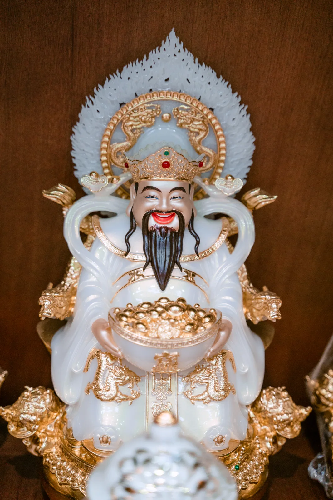 Tượng nhân vật râu đen đội mũ vàng, áo trắng trang trí vàng, hai tay ôm bát đầy hình thỏi tròn, phía sau có nền nhọn sáng và họa tiết rồng.