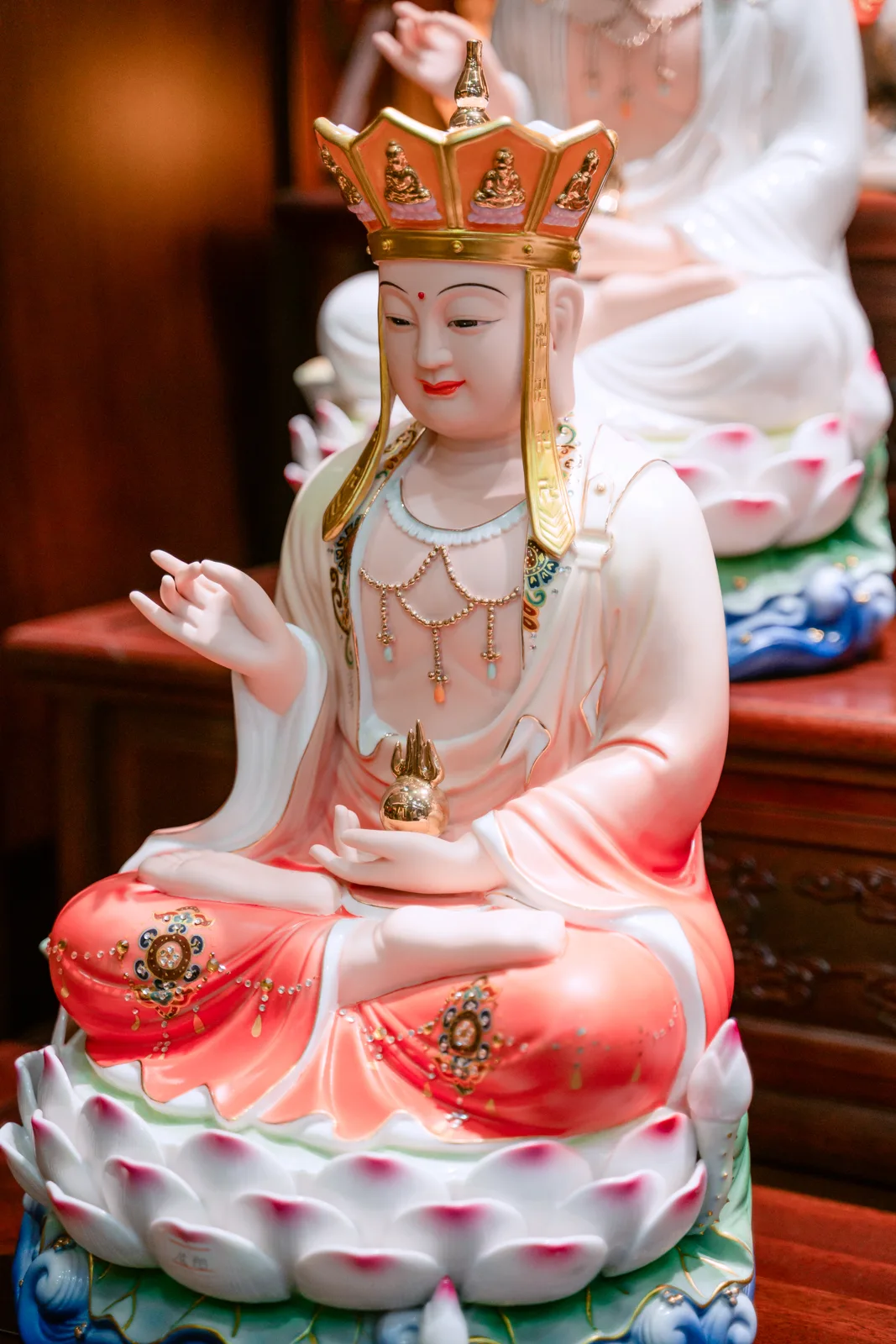 Tượng ngồi đội mũ cam nhiều cánh viền vàng, dải mũ rủ hai bên mặt, áo trắng chuyển đỏ, tay thấp nâng vật nhọn vàng trên đài sen.