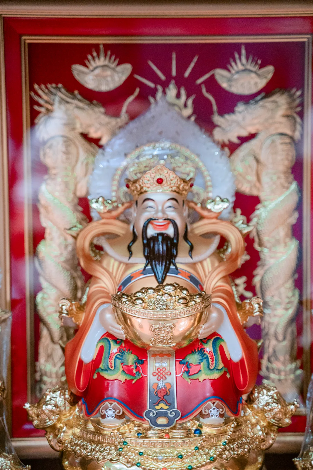 Tượng nhân vật râu đen đội mũ vàng, áo đỏ có họa tiết rồng xanh, hai tay ôm bát vàng lớn, phía sau là nền đỏ hình hai rồng.