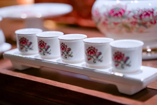 Năm chén trắng nhỏ họa tiết sen hồng xanh, miệng viền vàng, xếp một hàng trên khay trắng dài có chân thấp trên mặt nâu.